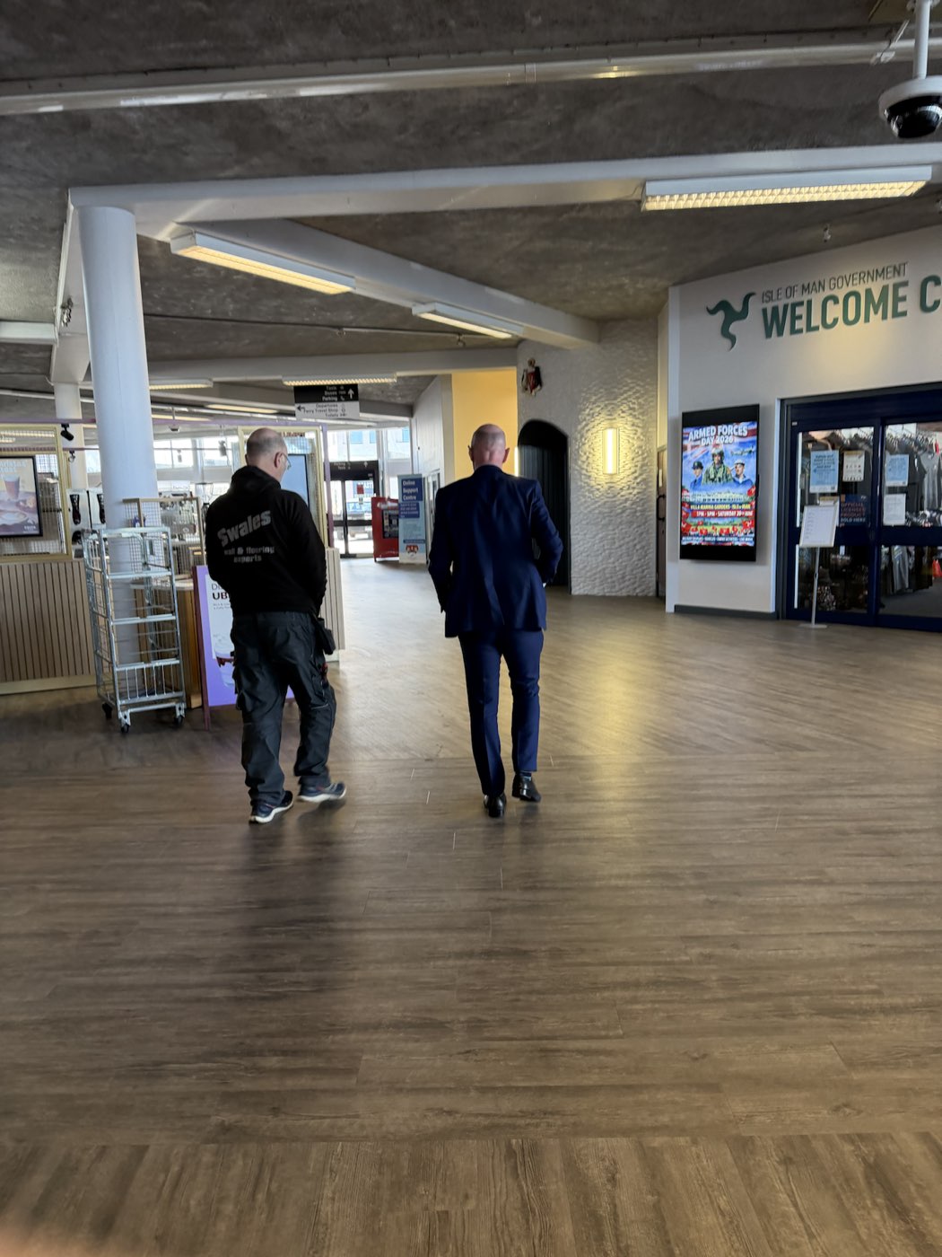 A Swales Flooring team member and a client walking across a newly fitted wood-effect floor in the bright open concourse of the Isle of Man Welcome Centre.