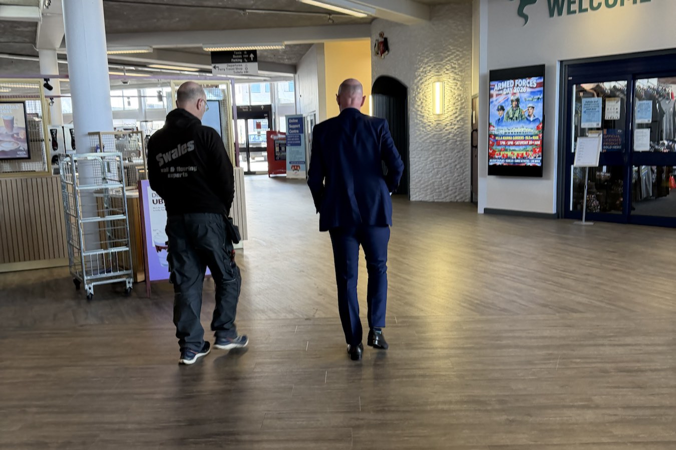 A wide view of a Swales Flooring team member and a client walking across a newly fitted wood-effect floor in the bright concourse of the Isle of Man Welcome Centre.